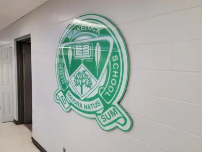 There is a green school crest made of a ceramic material hanging on the beige hallway wall