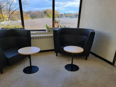 Small seating area shown in front of a window to the outside of the school. Two black curved comfortable chairs shown each with a small round wooden table in front of them. Each round wooden table top has a black pedestal bottom.