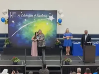 a stage set up at the front of a school gymnasium with one person speaking at a podium and two people standing centre stage. 